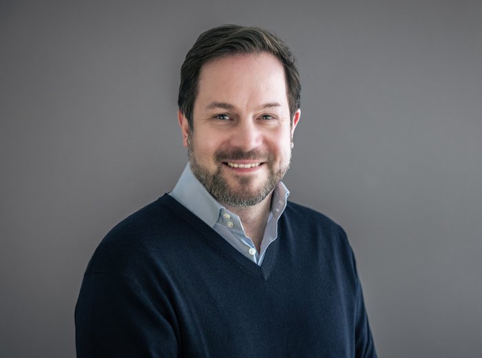 Porträt eines lächelnden Mannes, Bjoern Drews, mit kurzem dunklem Haar und Bart. Er trägt ein dunkles Oberteil mit einem hellen Hemdkragen darunter.