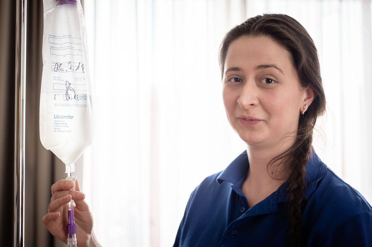 Eine Frau mit dunklen Haaren, die zu einem Zopf geflochten sind, trägt ein dunkelblaues Pflegeoutfit. Sie hält eine Infusionsflasche in der Hand und blickt ruhig in die Kamera.