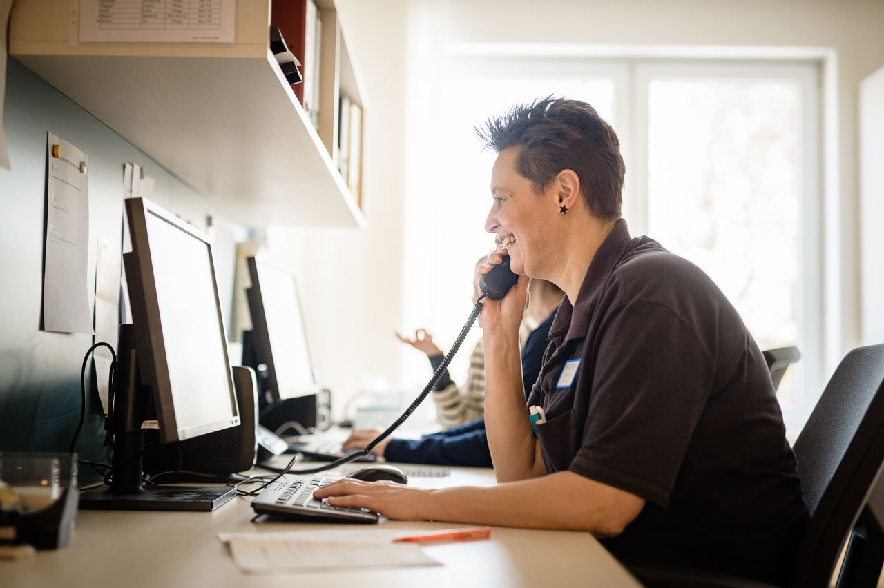 Eine Frau sitzt an einem Schreibtisch in einem modernen Büro. Sie trägt ein dunkles Poloshirt und spricht konzentriert in ein Telefon, während sie auf mehrere Computerbildschirme schaut. Das Büro ist hell, mit großen Fenstern, die Tageslicht hereinlassen.