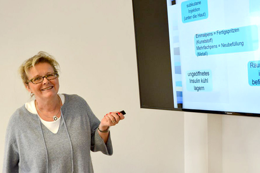 Das Bild zeigt eine Person, die eine Präsentation hält. Die Person steht vor einer Projektionsfläche oder einem Bildschirm, auf dem grafische Inhalte oder Folien angezeigt werden. Sie trägt eine Brille und ein legeres Outfit und gestikuliert mit einer Hand in Richtung der Präsentation.